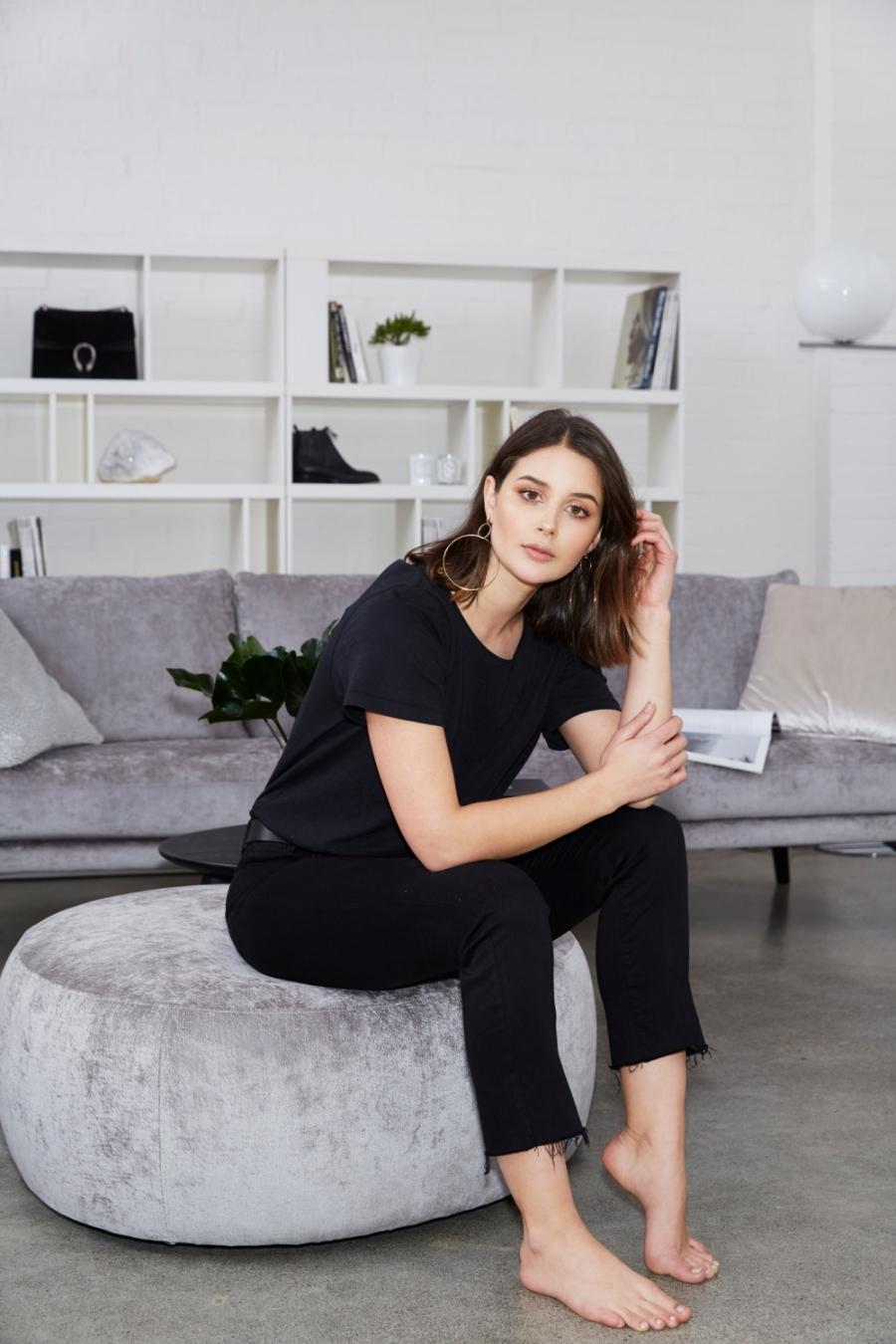 A woman in a black T-shirt and black jeans sits on a grey pouffe in a light-filled interior