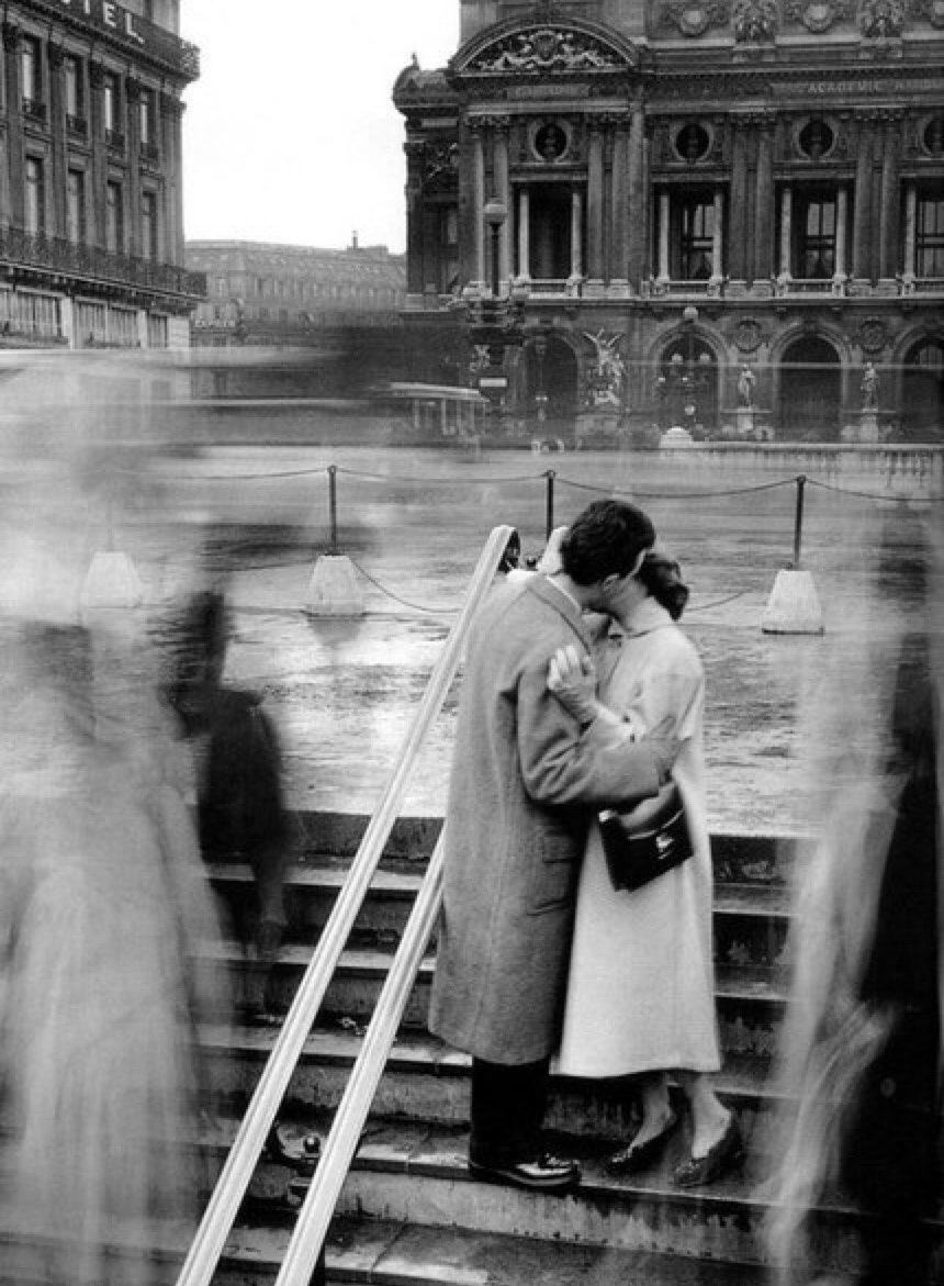 A couple in love kiss on the steps outside the Paris Opéra, passers-by blurred in motion