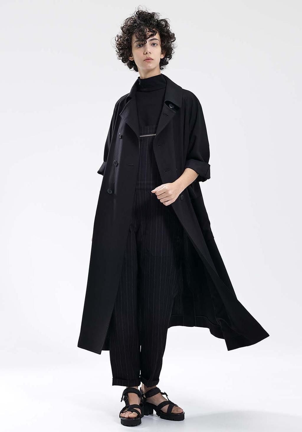 A model in a black trench coat worn over a black pinstripe jumpsuit