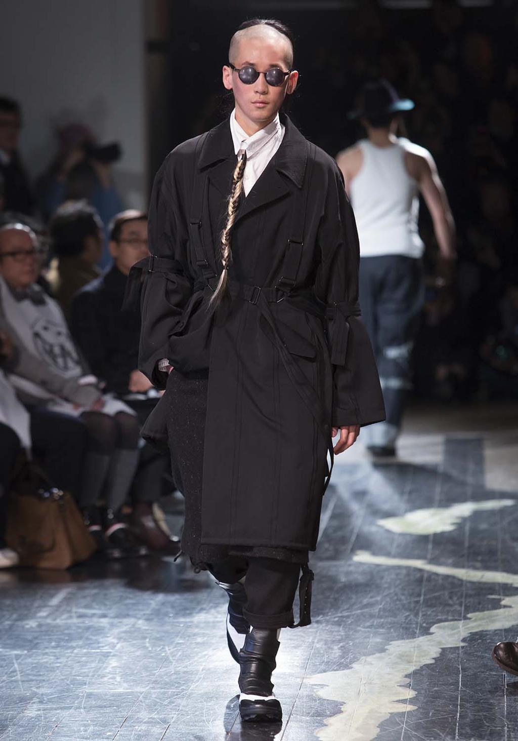 A man in a black belted trench coat with a long braid, on the runway