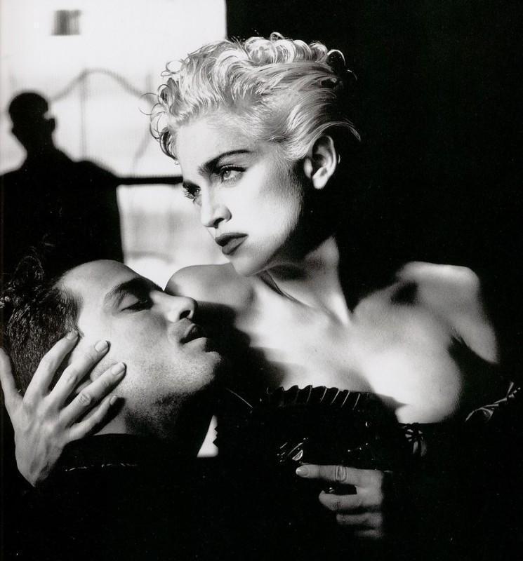 A fair-haired woman in a black corset holds the face of a reclining man — a high-contrast portrait