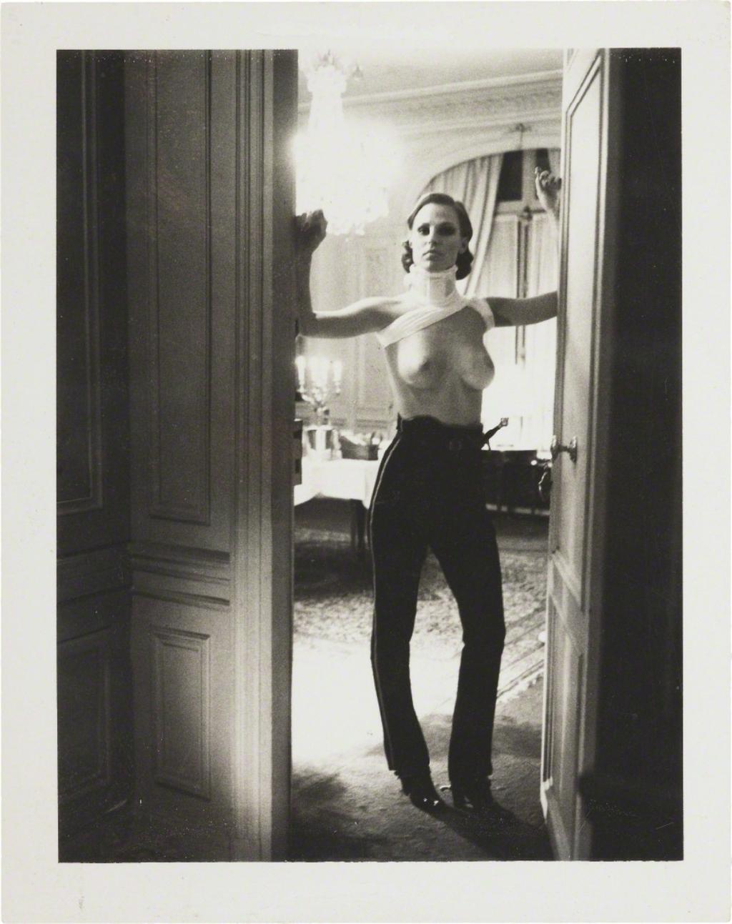 A semi-nude woman in black trousers stands in the doorway of a lavish dining room