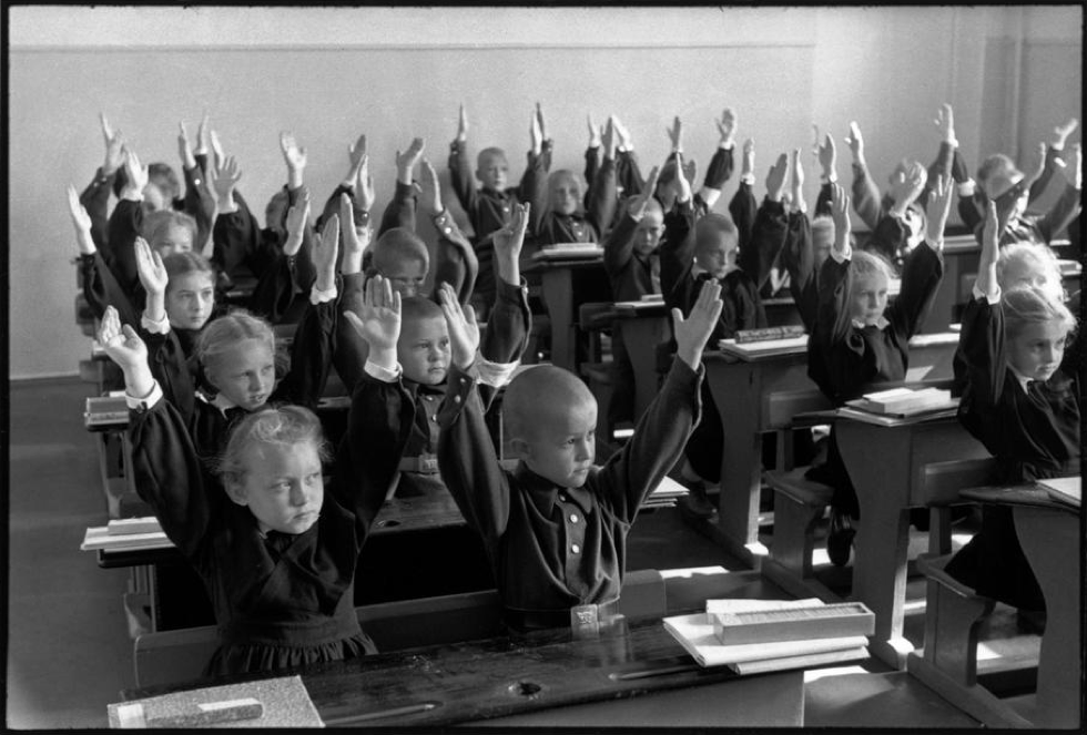 Schoolchildren sit at their desks and raise their hands in unison in the classroom