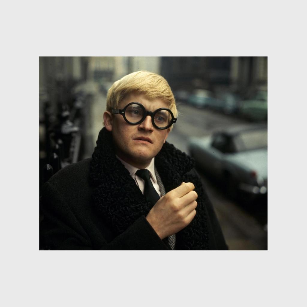 A young David Hockney in round glasses and a black coat on a city street