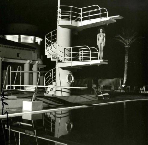A fair-haired man in swimming trunks wearing a monocle stands beside a pool with swimmers
