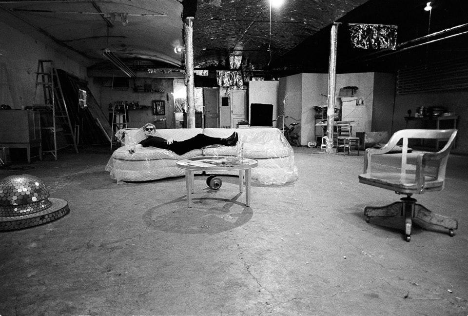 Black-and-white photograph of the Factory studio: Warhol lying on a sofa in a large room with a concrete floor