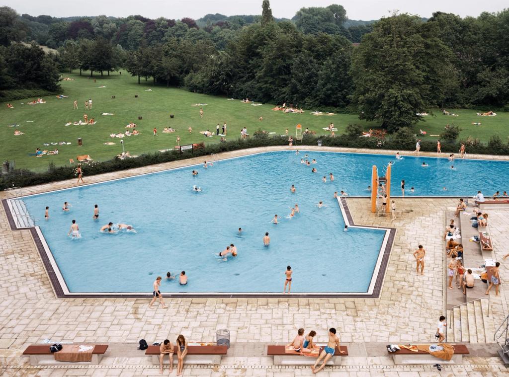 An open-air swimming pool with bathers, a sunbathing lawn and trees alongside