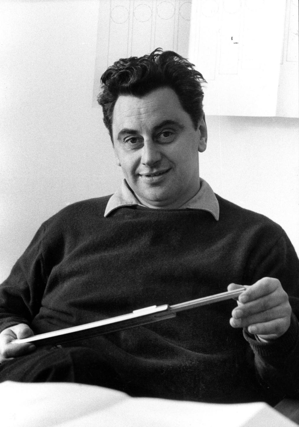 A black-and-white portrait of a young designer in a sweater, holding a slender metal component