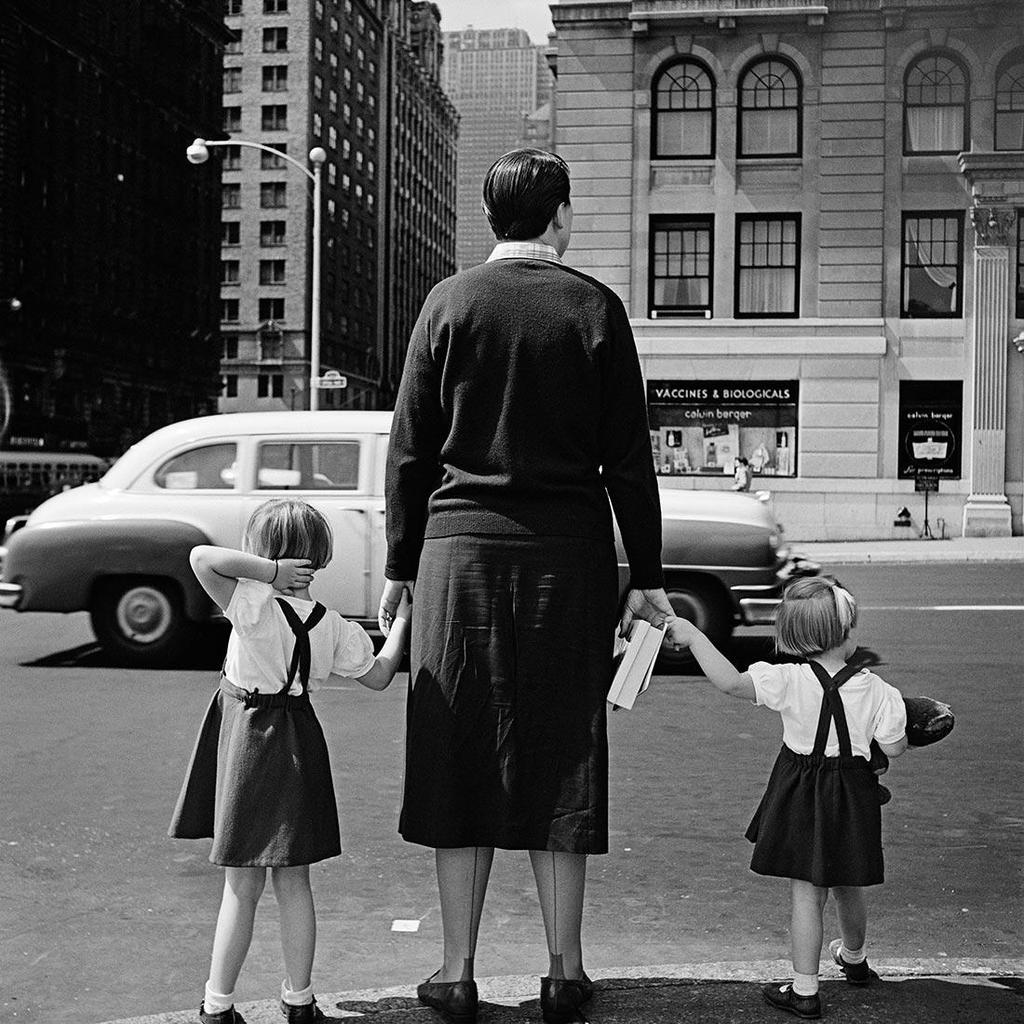 Black-and-white photograph from behind: a woman holds the hands of two girls at the edge of a city road