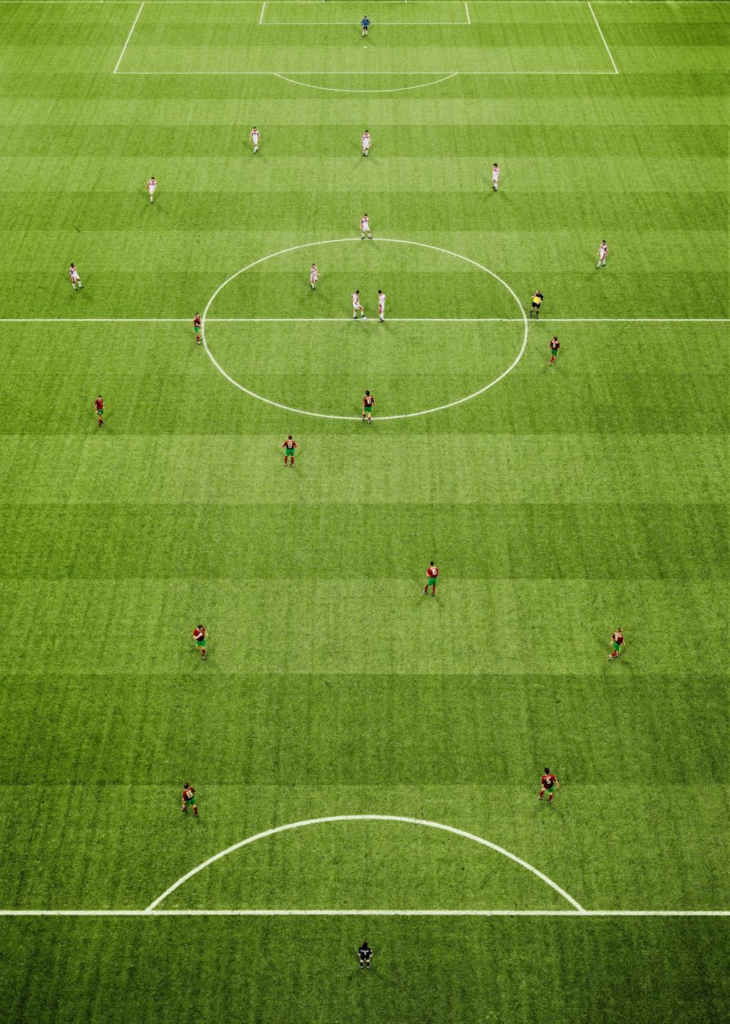 A football pitch from above: symmetrical markings and players from two teams on green turf