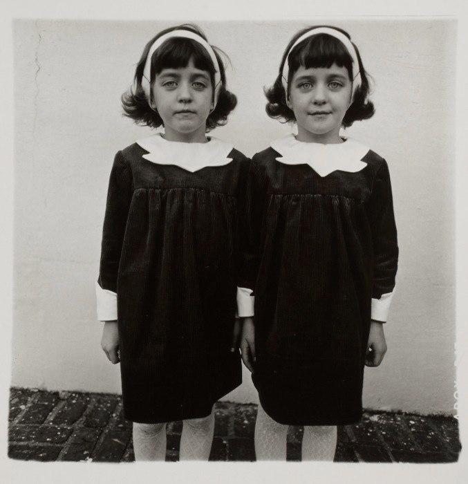 A black-and-white portrait of twin girls in identical dark dresses with white collars, by Diane Arbus