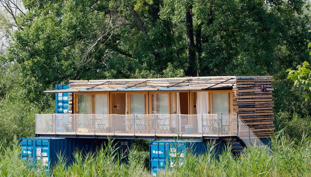 Длинный фасад Containhotel из синих контейнеров с террасой среди деревьев