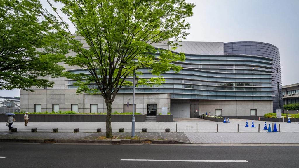 The Kyoto Concert Hall seen through a row of trees, its curved ribbon facade with horizontal glazing