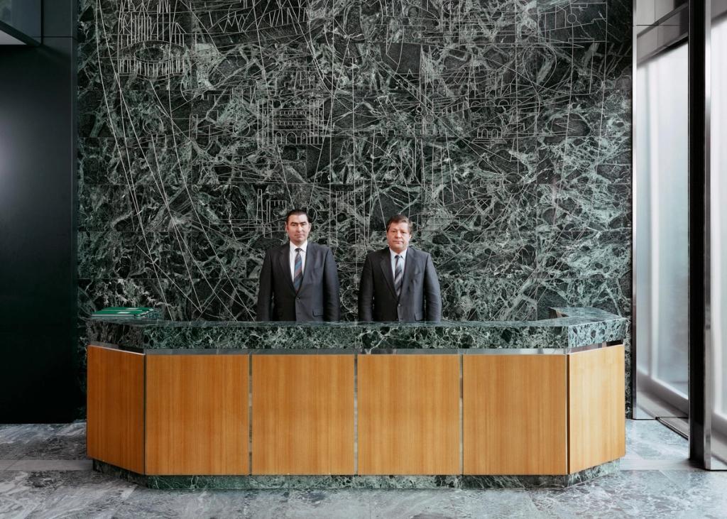 Two men in suits at a reception desk against a wall of green marble