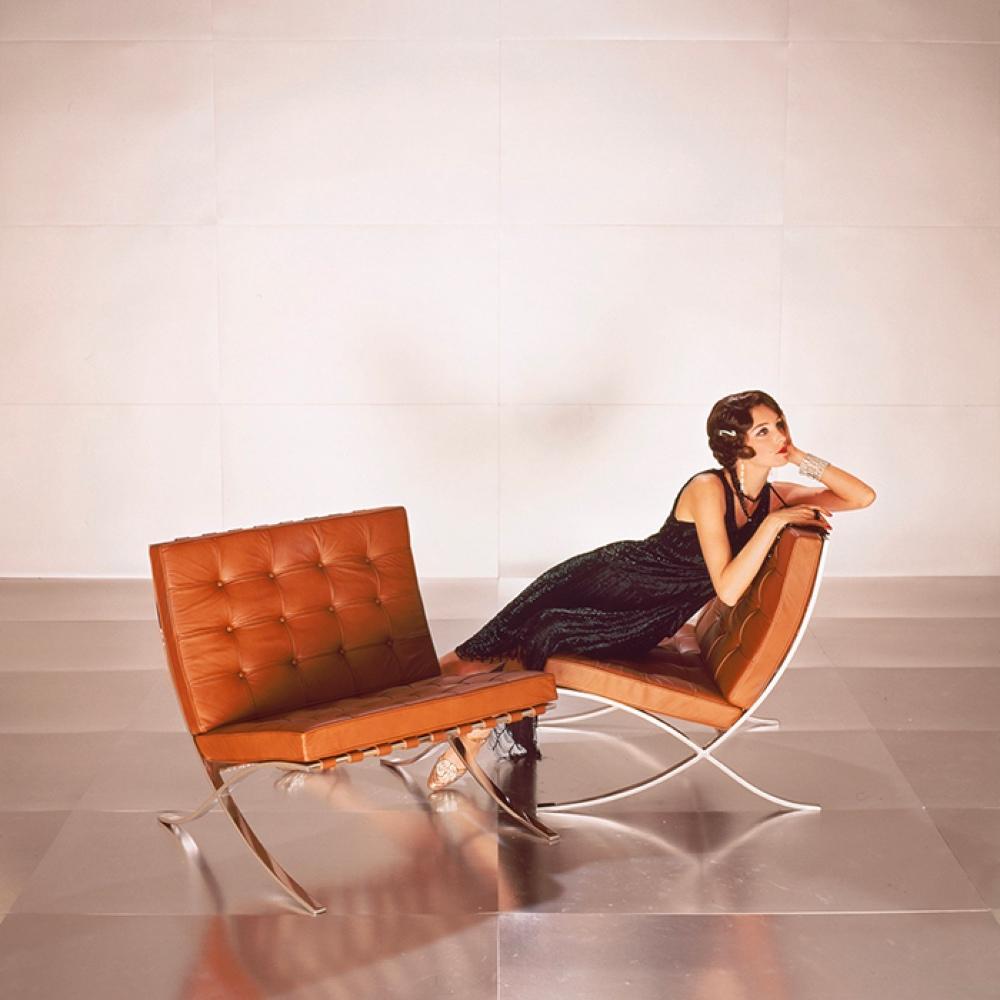 A woman in a black dress reclines across Barcelona Chairs upholstered in tan leather