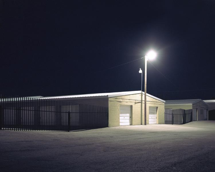 Storage units behind a metal fence at night, lit by the harsh glow of a solitary streetlamp