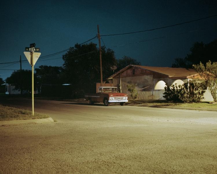 An old pickup truck on a suburban street at night beside an 11th St road sign under a twilight sky