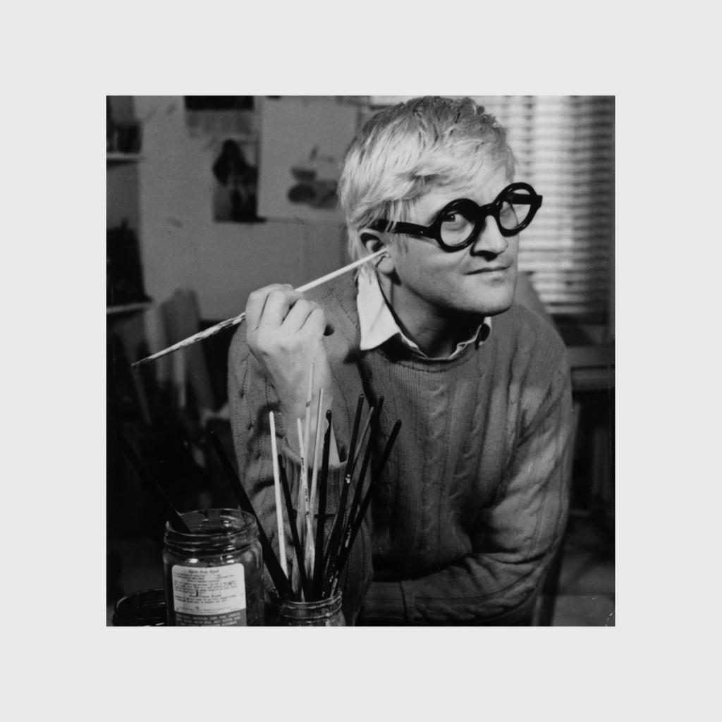 A black-and-white shot of a young Hockney holding a brush in his studio