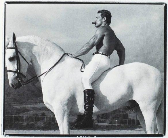 A muscular man with a cigar on horseback, riding a white horse against a backdrop of hills