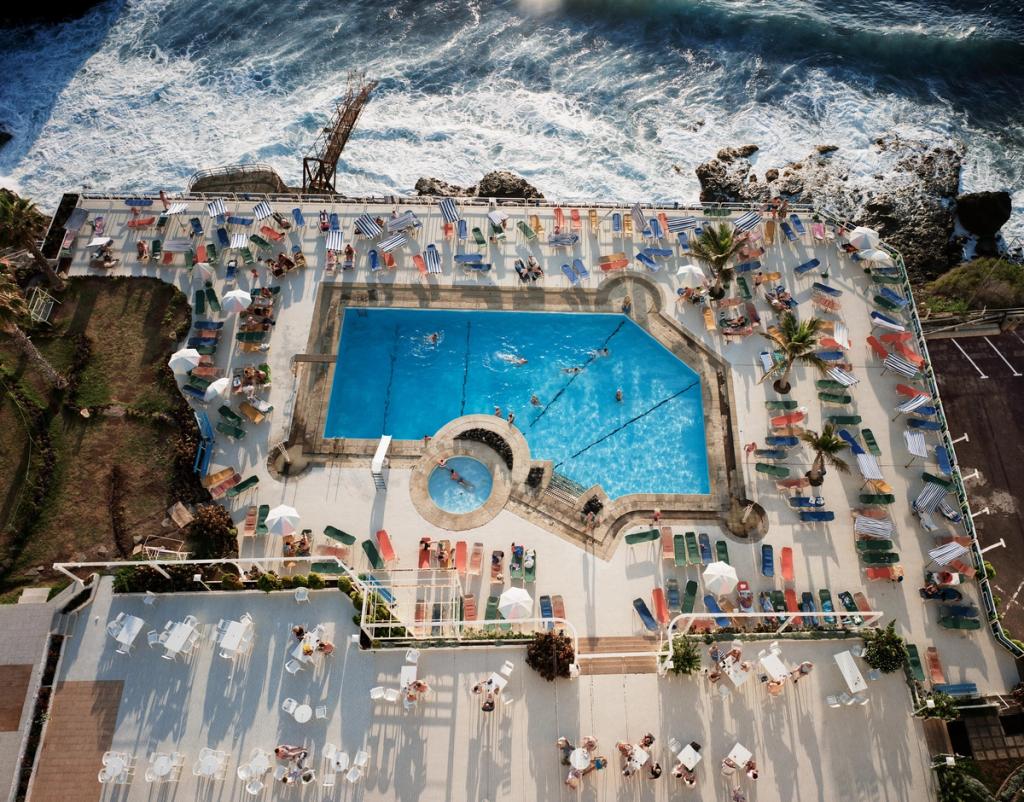 An aerial view of a blue swimming pool with sun loungers on a terrace beside a churning ocean