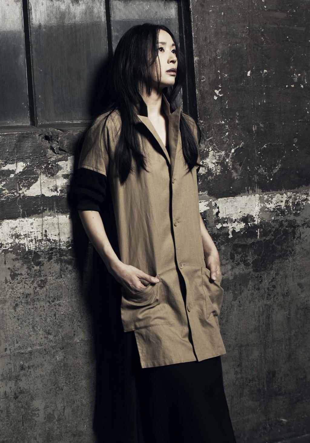 A woman in a loose khaki shirt against a weathered wall, black-and-white photograph