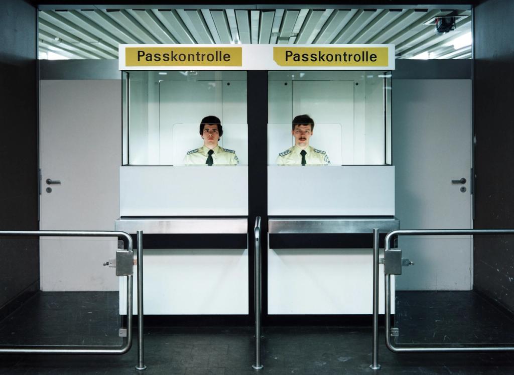 Two border guards in identical passport-control booths marked Passkontrolle