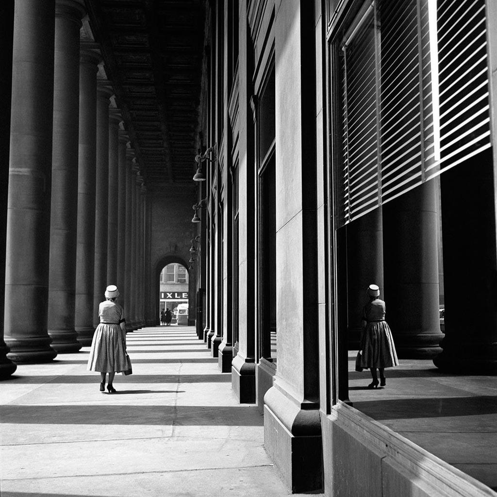 Black-and-white photograph: a woman in a dress and hat walks along a colonnade, her reflection visible in a shop window
