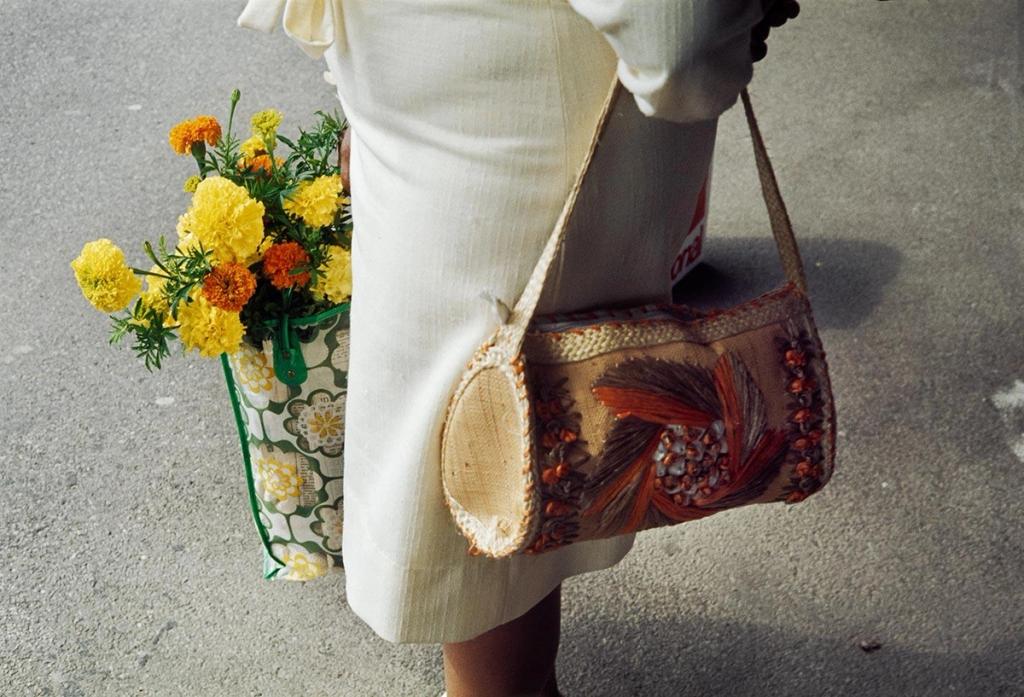 Colour photograph: a woman in a cream dress carrying a bouquet of marigolds and a woven shoulder bag