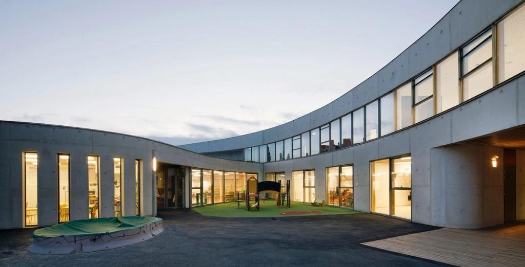 The concrete curvilinear courtyard of Ecole Jean-Moulin school, with a play area and illuminated windows