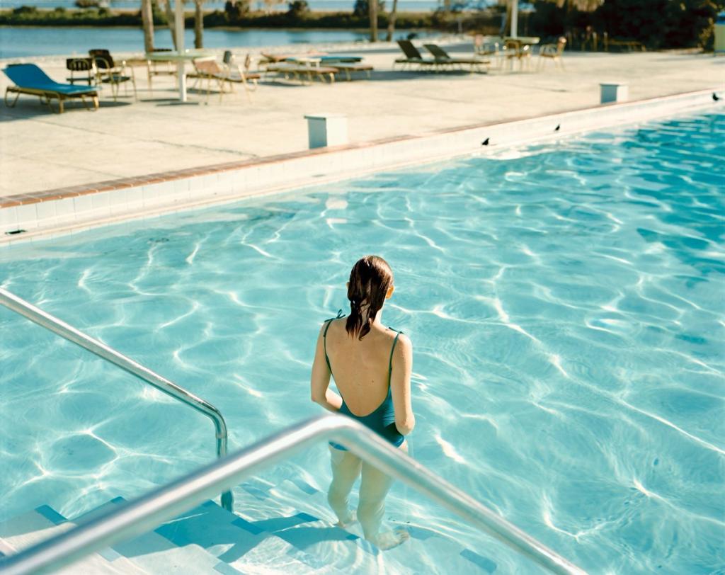 A colour photograph by Stephen Shore: a woman in a swimsuit stands in the turquoise water of a swimming pool, with sun loungers on the poolside