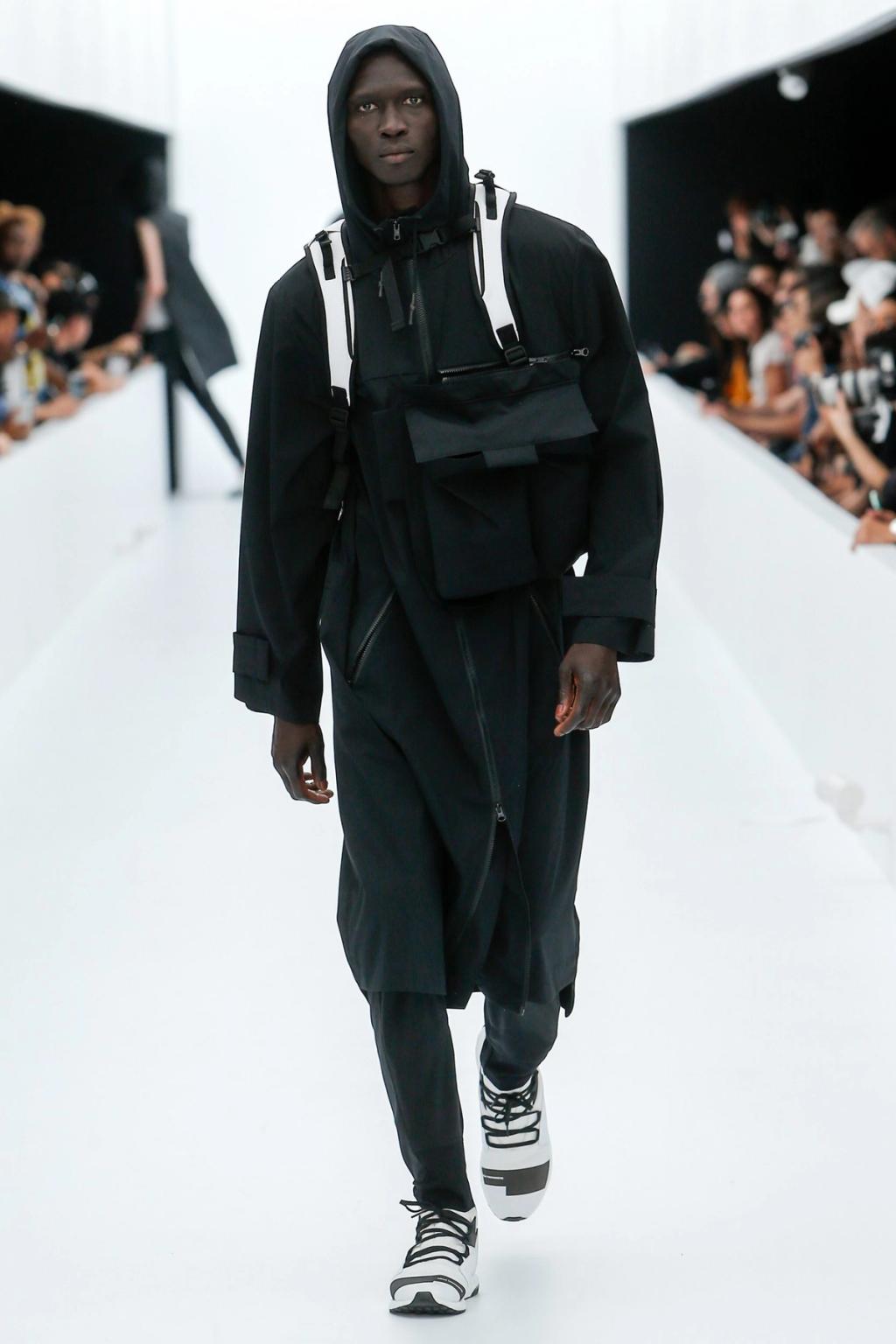 A Y-3 model in a black jacket with a chest bag and trainers on the runway