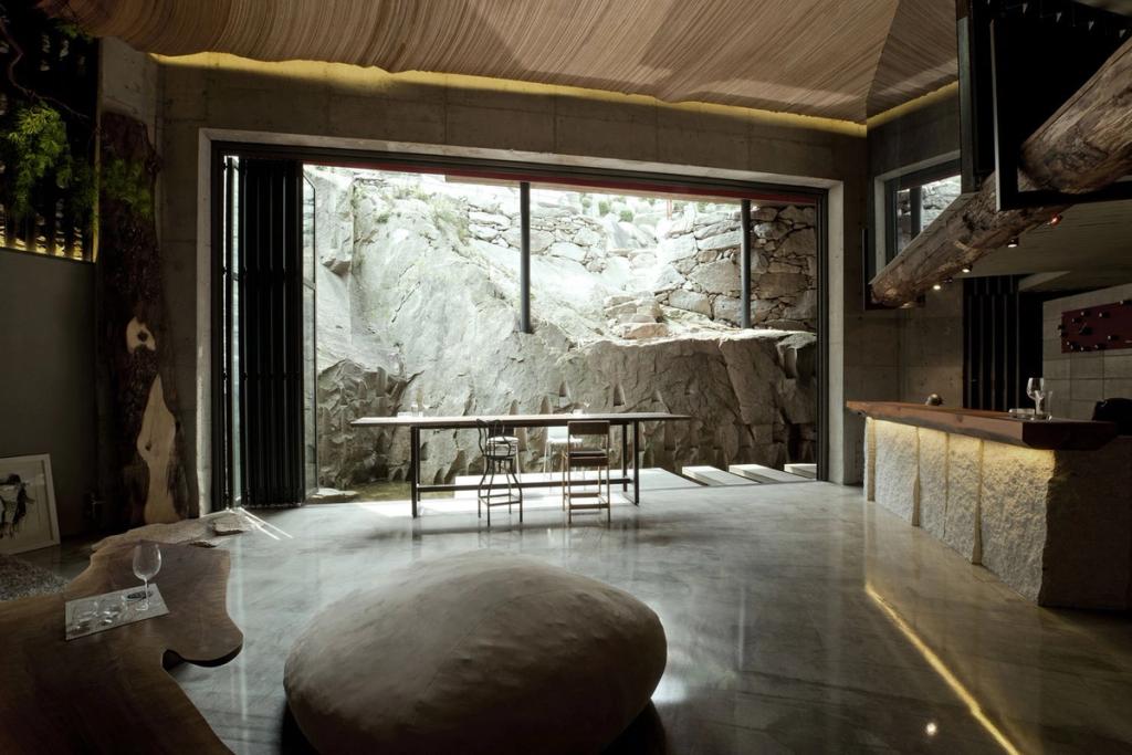 A living area with a sliding glass wall opening onto a stone rock face, a wooden bench and a boulder used as a pouf