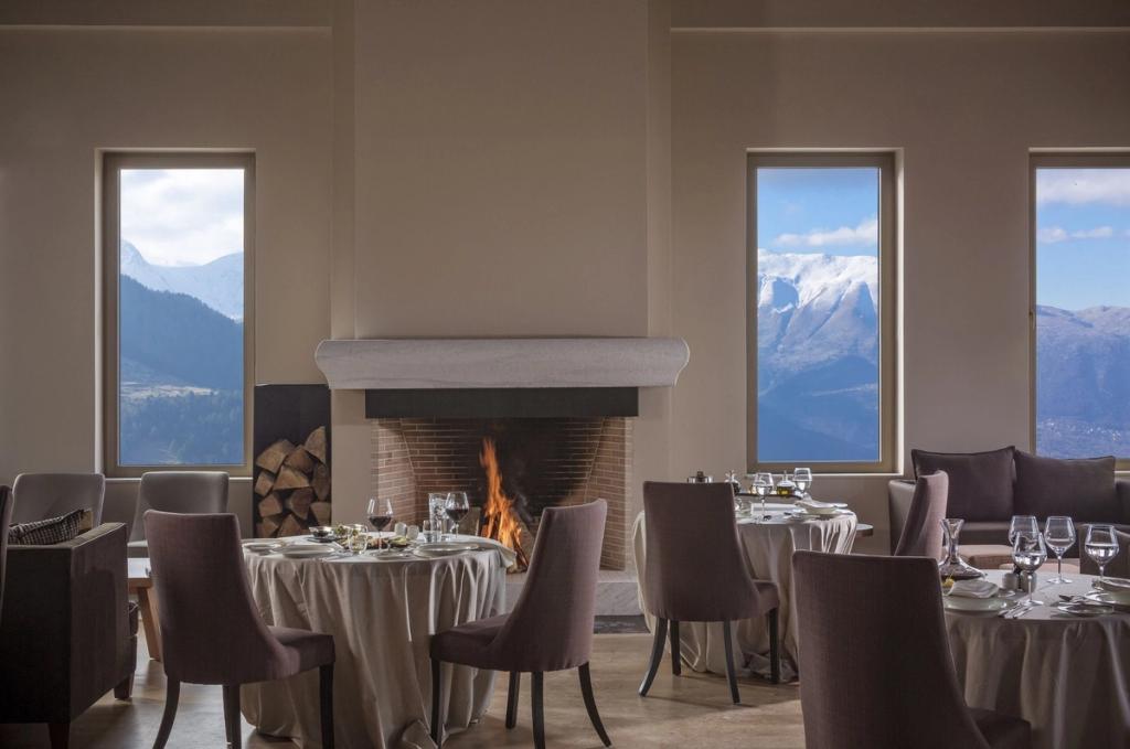 A restaurant with a burning fireplace and windows overlooking the snow-covered Pindus mountains