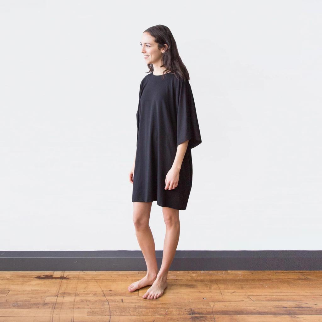 A model in a black oversized Encircled dress stands barefoot on a wooden floor