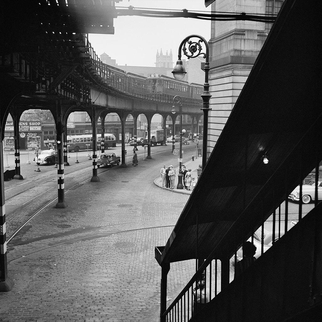 Black-and-white photograph: an elevated railway and a tram above a cobbled street, seen from a staircase