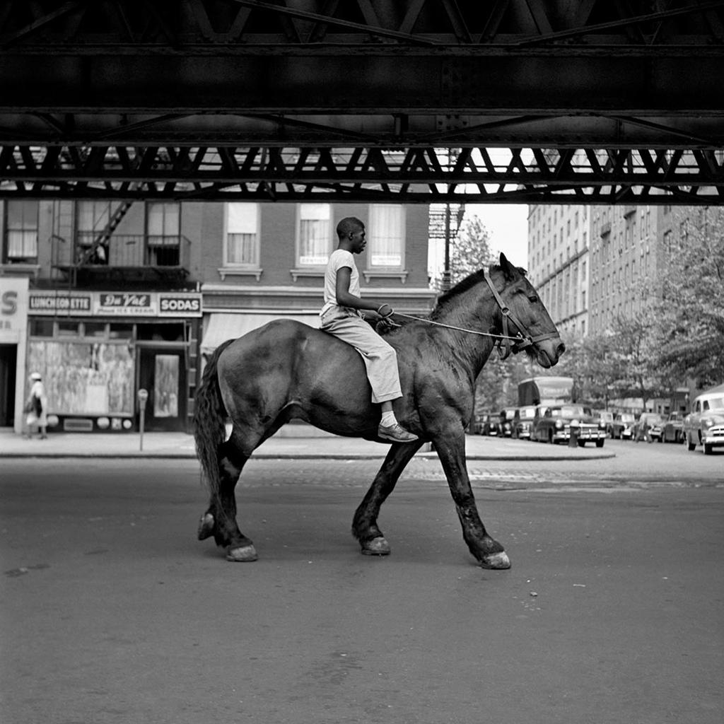 A self-portrait of Vivian Maier reflected in a mirror being carried by a mover, with houses in the background