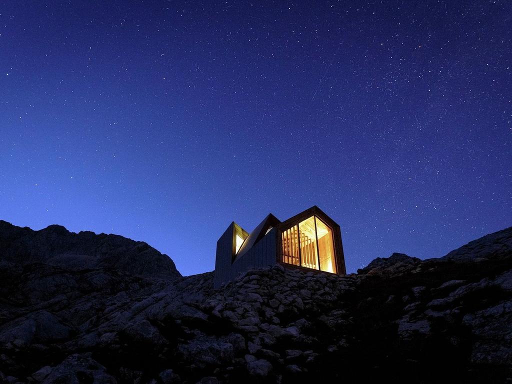 Силуэт бивуака Alpine Shelter Skuta со светящимися окнами под звёздным ночным небом в горах