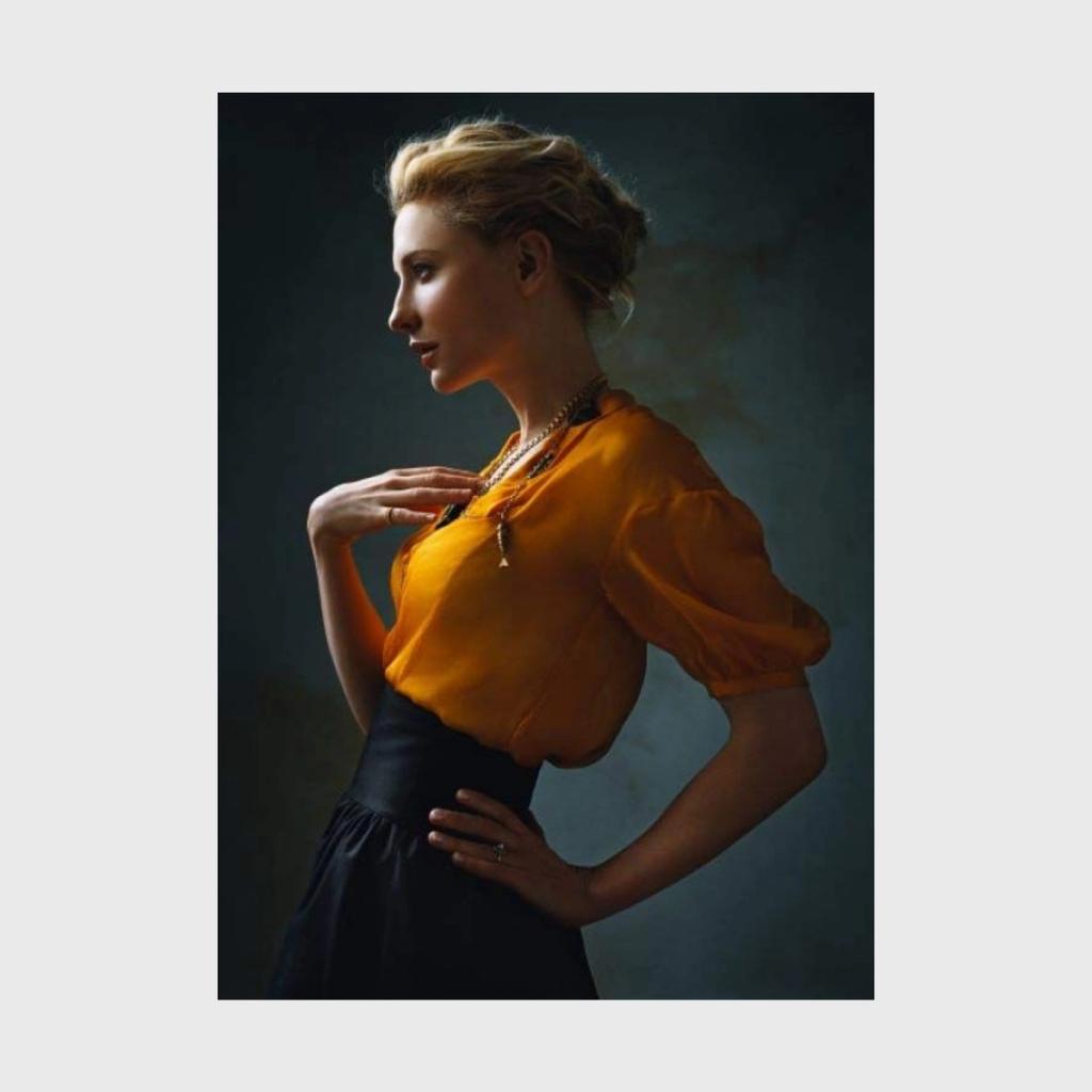 A colour profile portrait of a blonde woman in an orange blouse and black skirt against a dark background