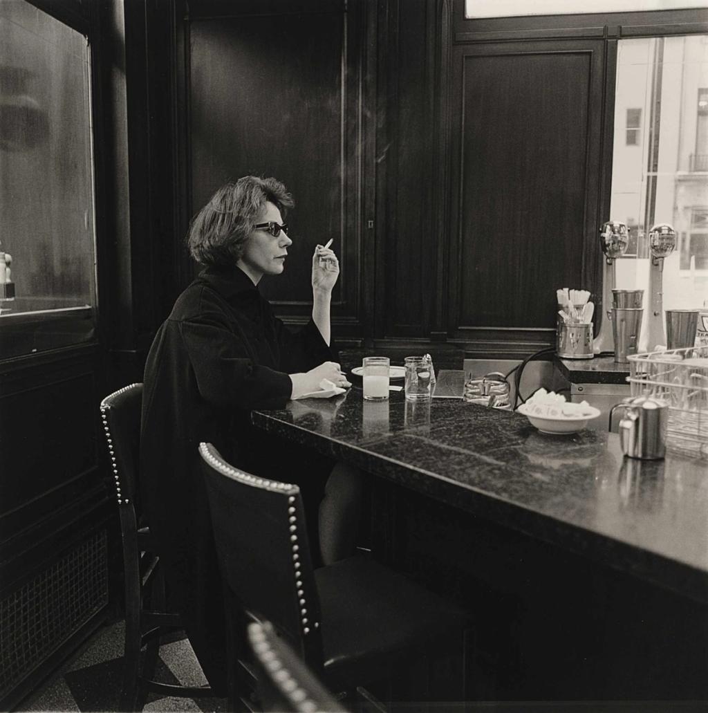 A woman in a dark coat and glasses smoking a cigarette at the counter of an empty café