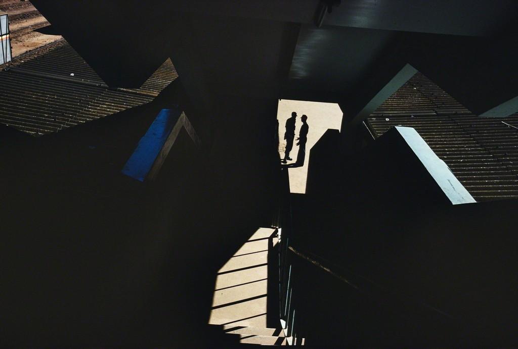 Two silhouettes caught in a pool of light amid dark geometric staircases and concrete structures