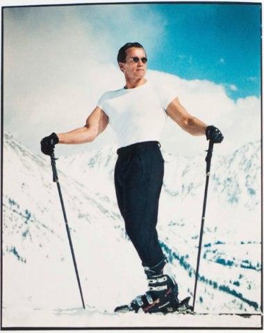 A man in a white T-shirt stands on skis against a backdrop of snow-covered mountains