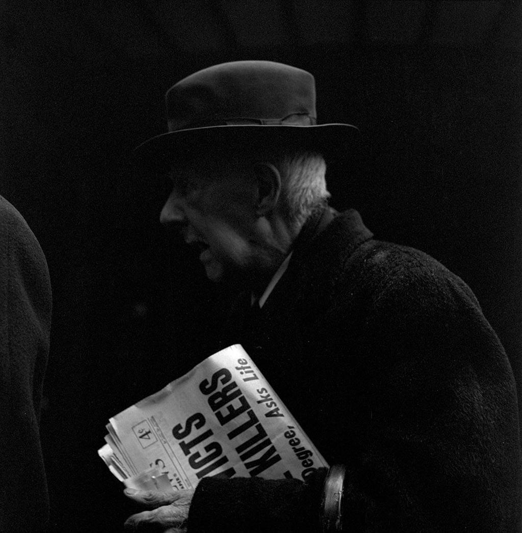 Black-and-white photograph: an elderly man in a hat carrying a newspaper steps out of shadow into light