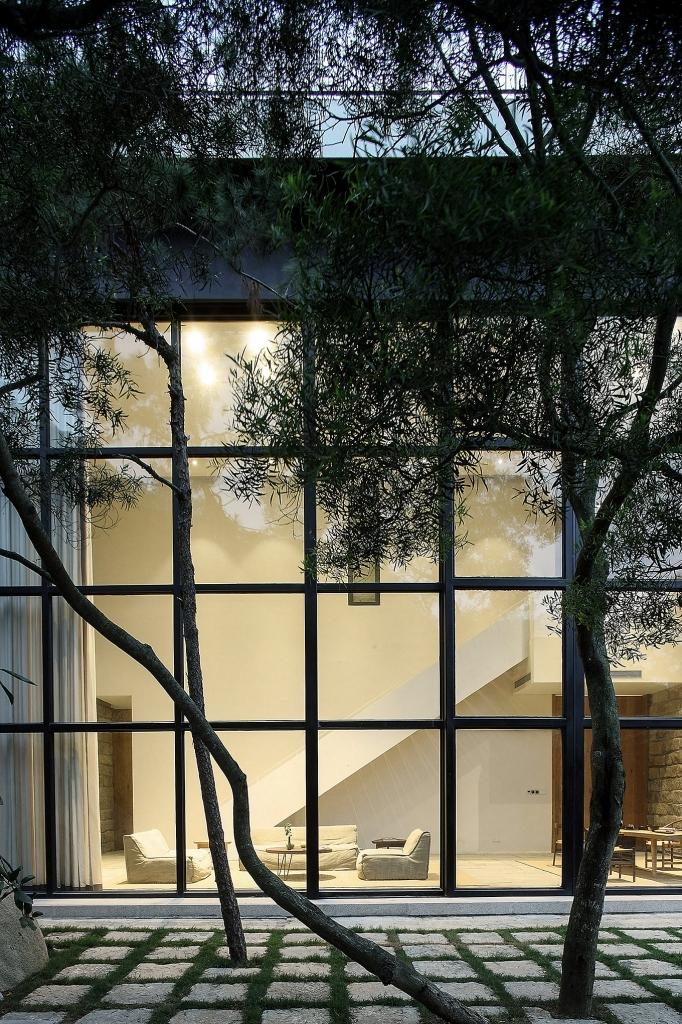 The double-height façade of a house with a glass wall glimpsed through olive trees at dusk
