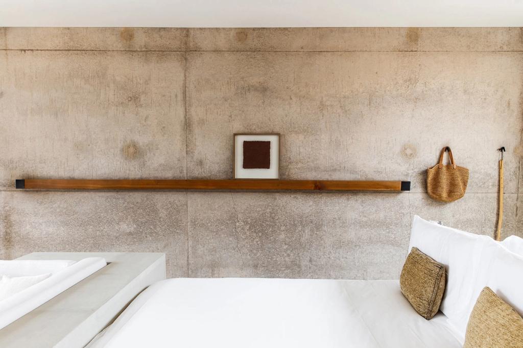 An Amangiri bedroom with a white bed, a wooden shelf and a woven bag against a concrete wall