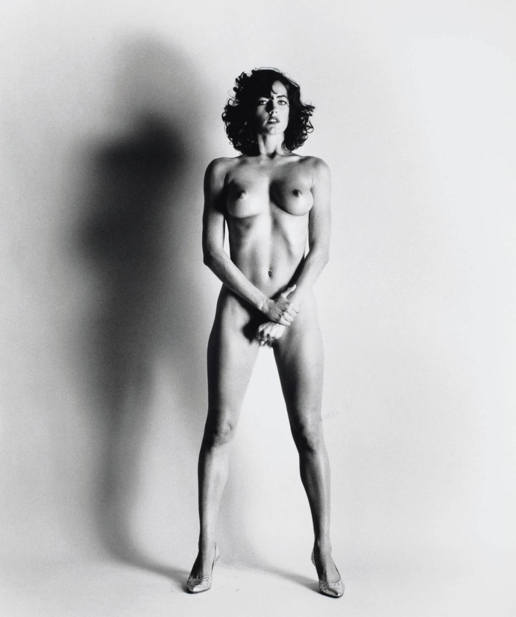 A nude woman in heeled shoes stands full-length against a pale wall, her shadow cast beside her