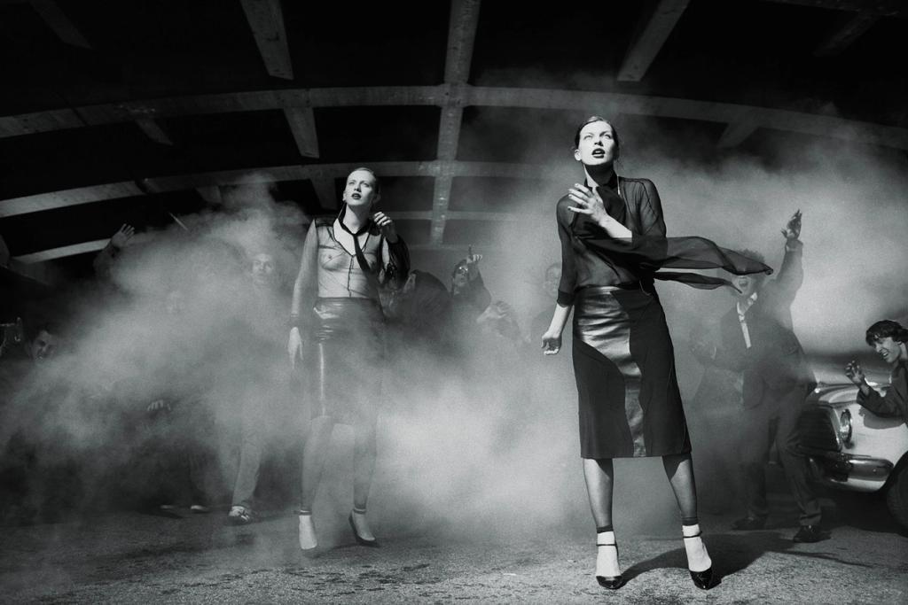 A black-and-white scene: two models among smoke and people with raised hands in a garage