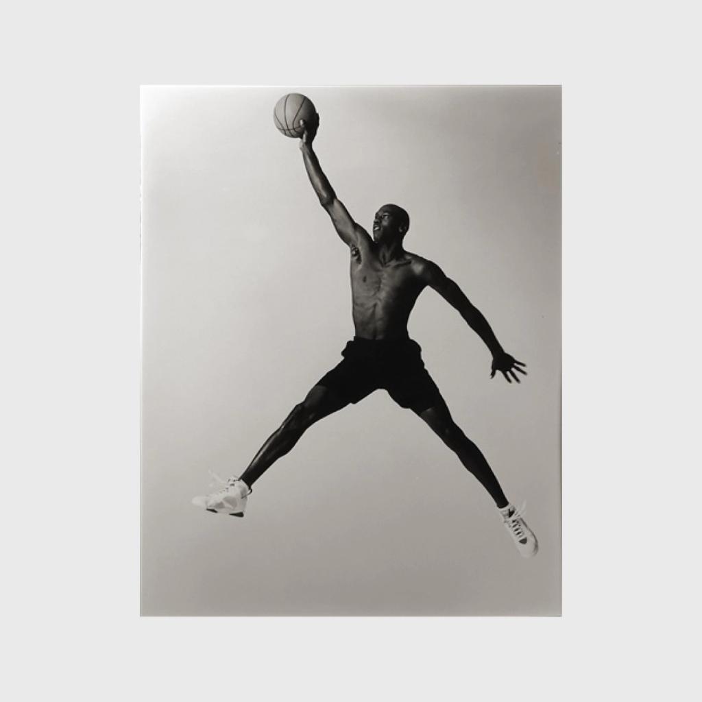 A black-and-white photograph of a basketball player mid-jump, one arm raised above his head, ball in hand