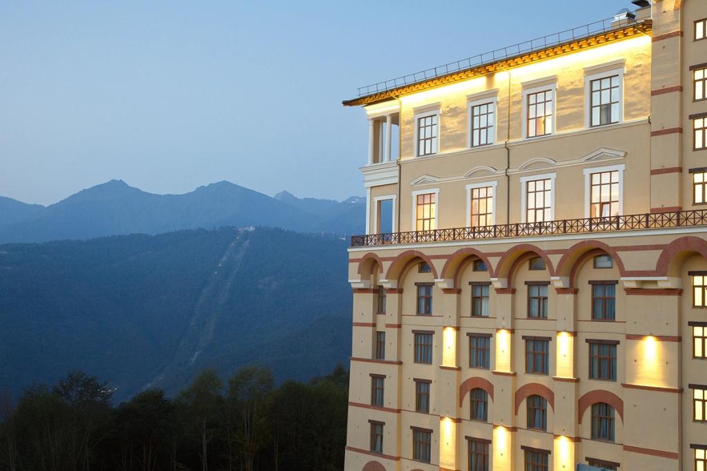 The illuminated façade of Solis Sochi Hotel at dusk, with the mountain ridge in the background
