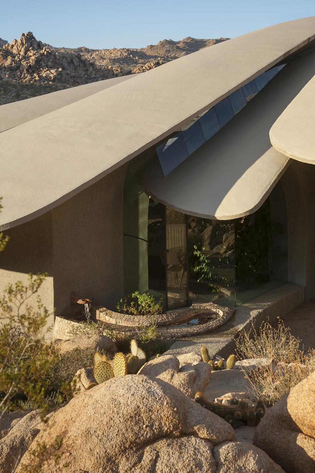 The glass entrance of a house beneath concrete petal vaults, surrounded by cacti and granite boulders