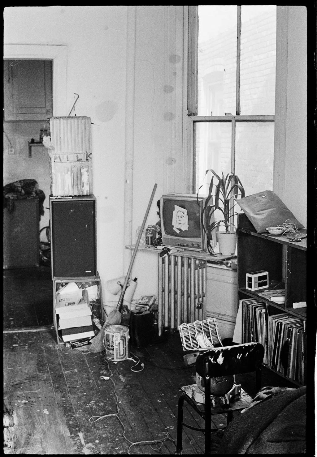 A black-and-white photograph of a cluttered studio: an old television, speakers, a broom, and stacks of canvases against the wall